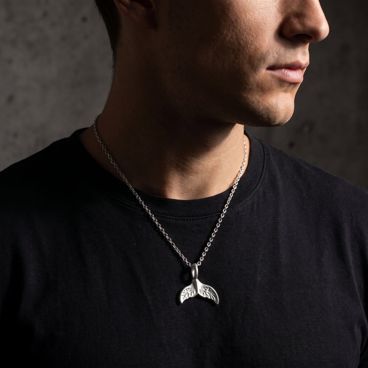 Right-facing profile of a man wearing a silver whale tail necklace over a black crew-neck shirt.