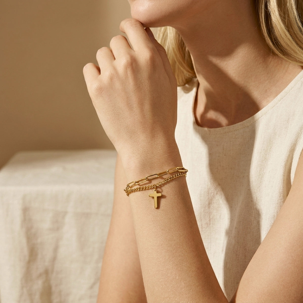 Woman wearing the gold-tone double-chain cross bracelet near her face with a white top in warm light.