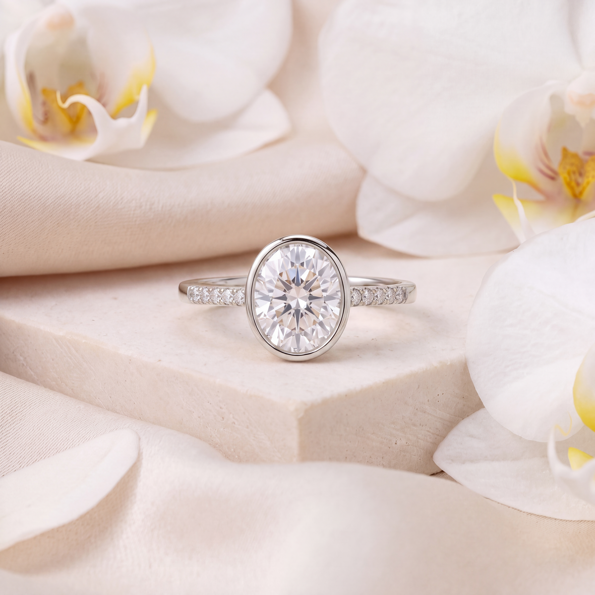 White gold oval moissanite bezel engagement ring with pavé side stones on a beige pedestal.