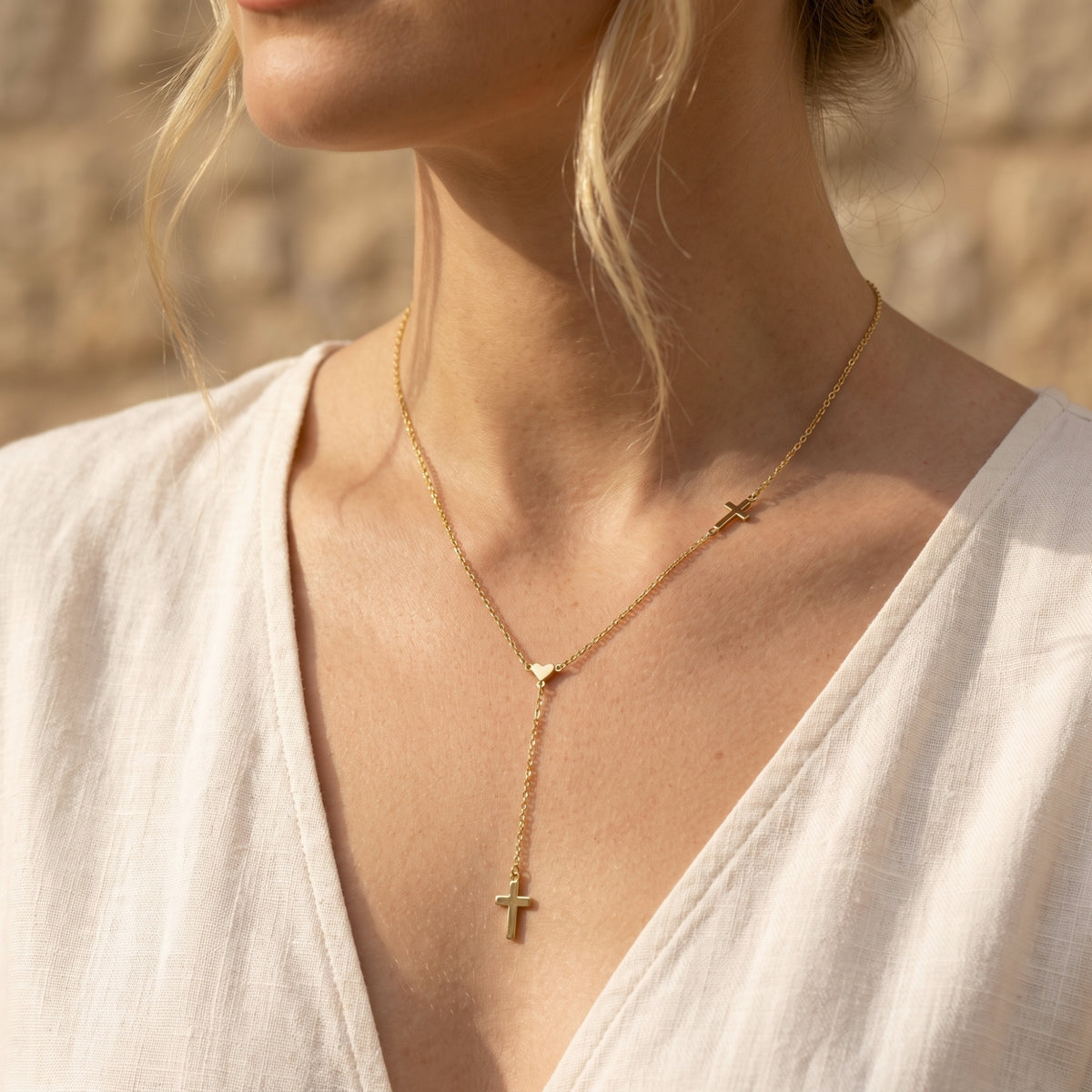Front view of a person wearing the gold-tone heart lariat necklace with a drop cross pendant over a white V-neck top.