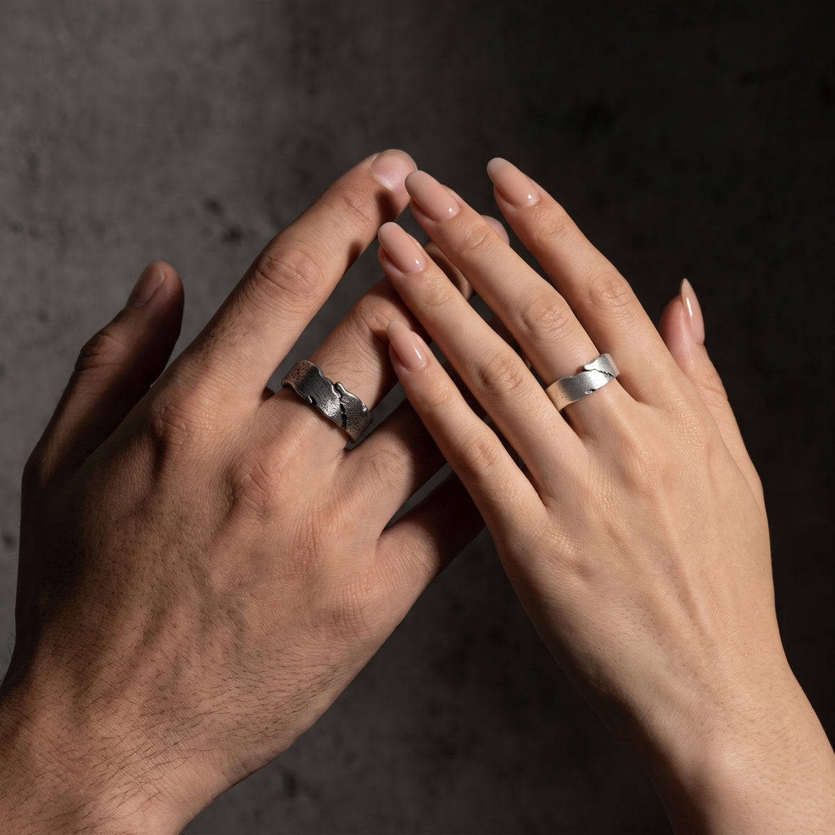 Two hands posed together against a dark textured background, each wearing a silver open fissure ring.