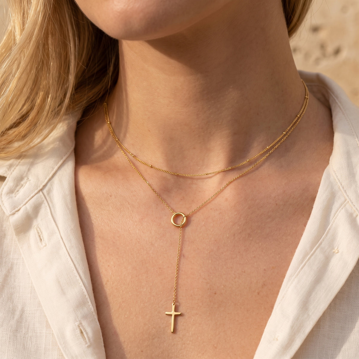 Gold-tone layered lariat cross necklace styled on a model wearing an open white linen button-down shirt.