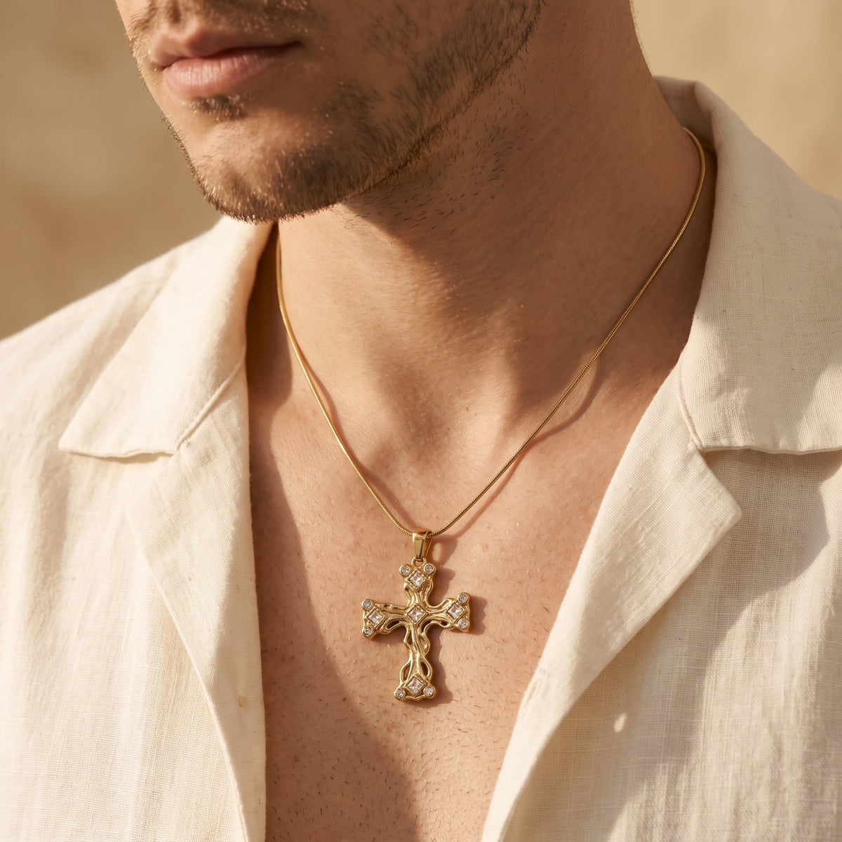 Man wearing a gold-tone filigree cross necklace with clear stone accents over an open-collar light shirt.