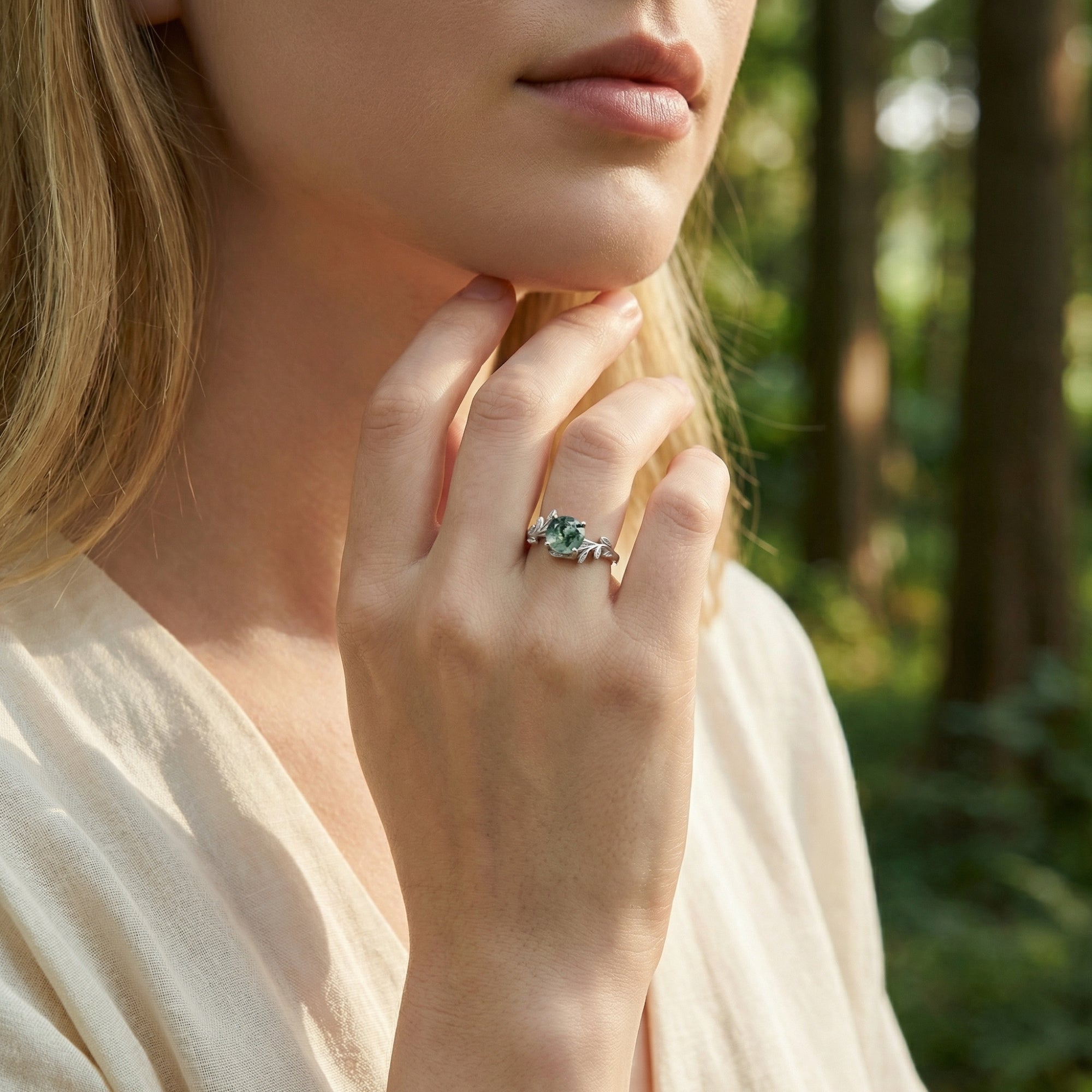 Silver floral crest moss agate ring worn on a model's hand against a natural forest background.