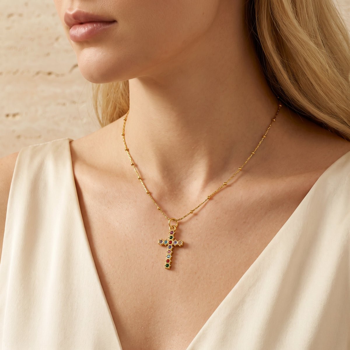 Gold-tone multicolored cross necklace on a model in a wider angled view with a cream V-neck garment.