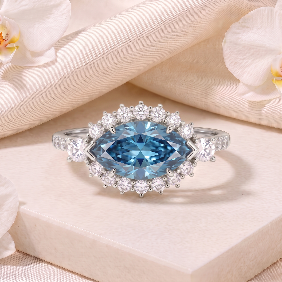 Front view of a white gold marquise blue moissanite halo ring resting on a cream pedestal.