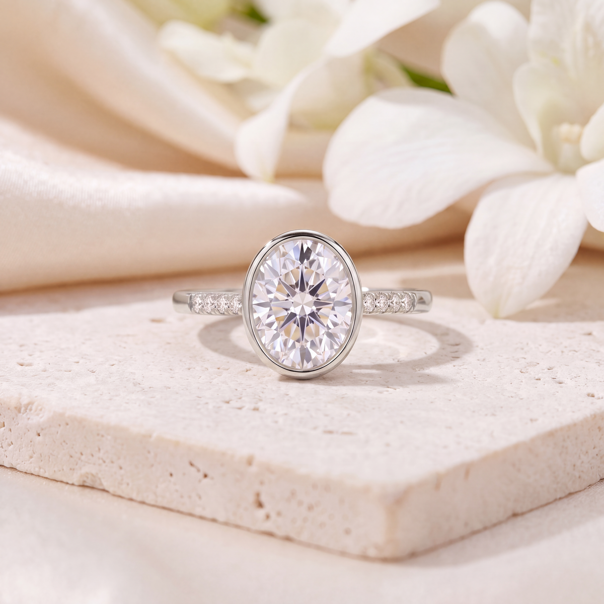 White gold oval moissanite bezel ring with pavé side stones on a beige stone block.