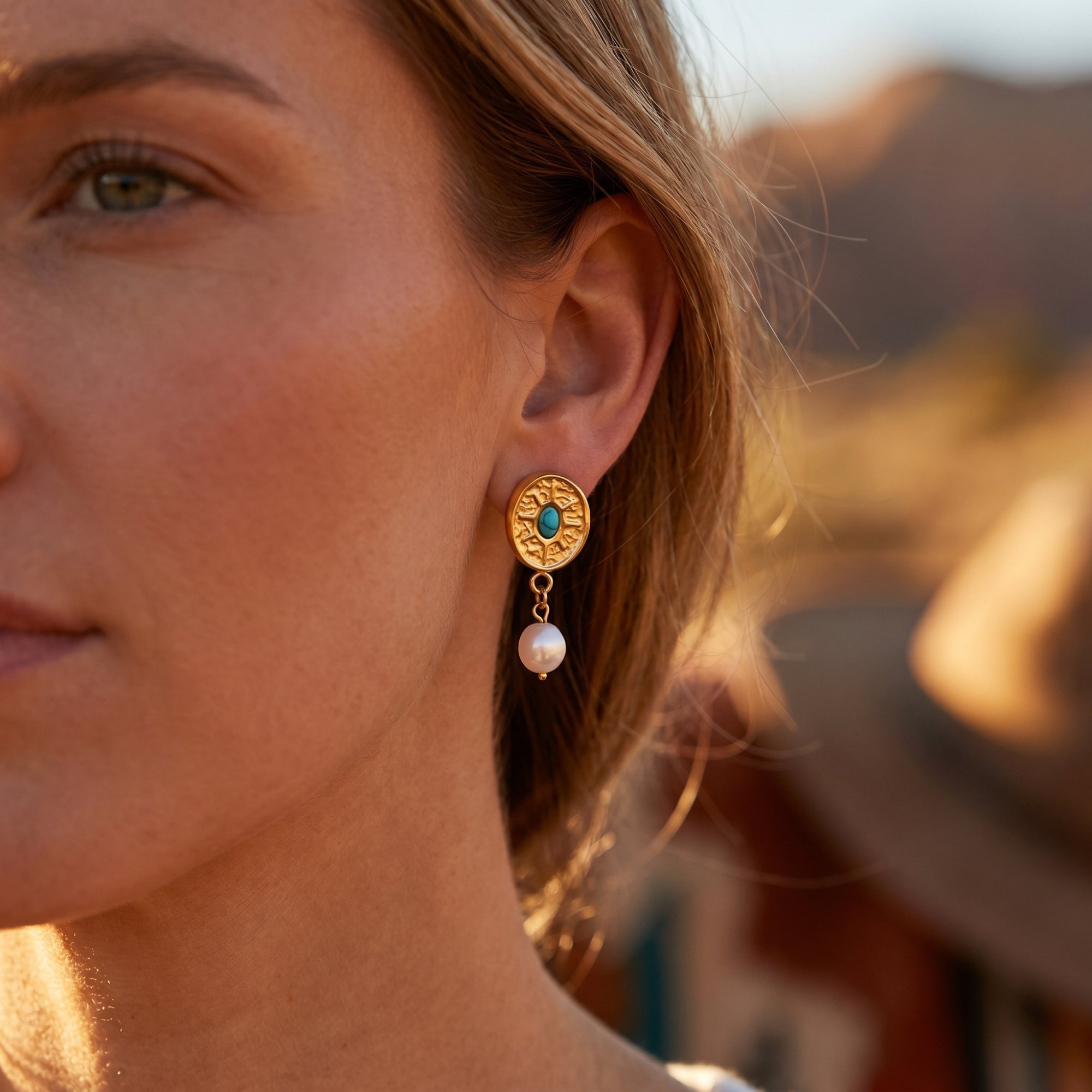 Woman wearing gold sunburst drop earrings with turquoise and pearl accents against a blurred desert background.