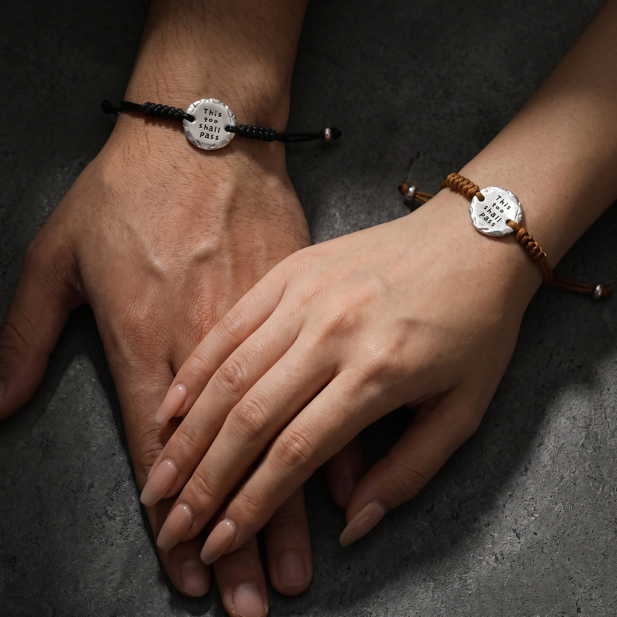 Two hands resting together, wearing matching brown and black cord bracelets with engraved silver medallions.