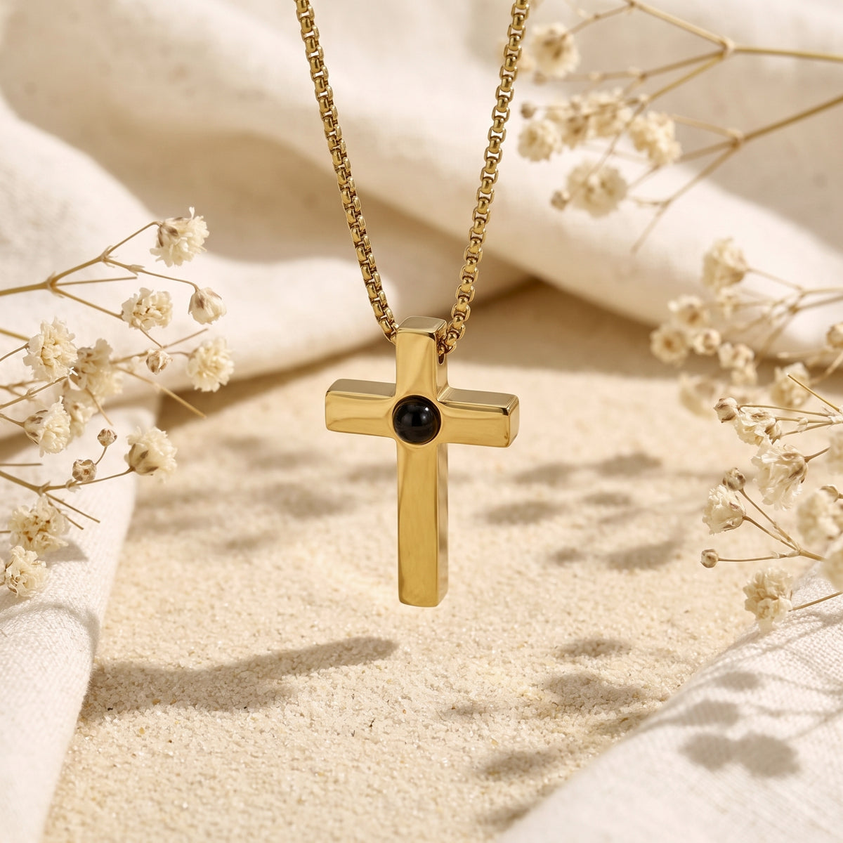 Gold-tone cross pendant with a central dark lens hanging above sand between dried white flowers.