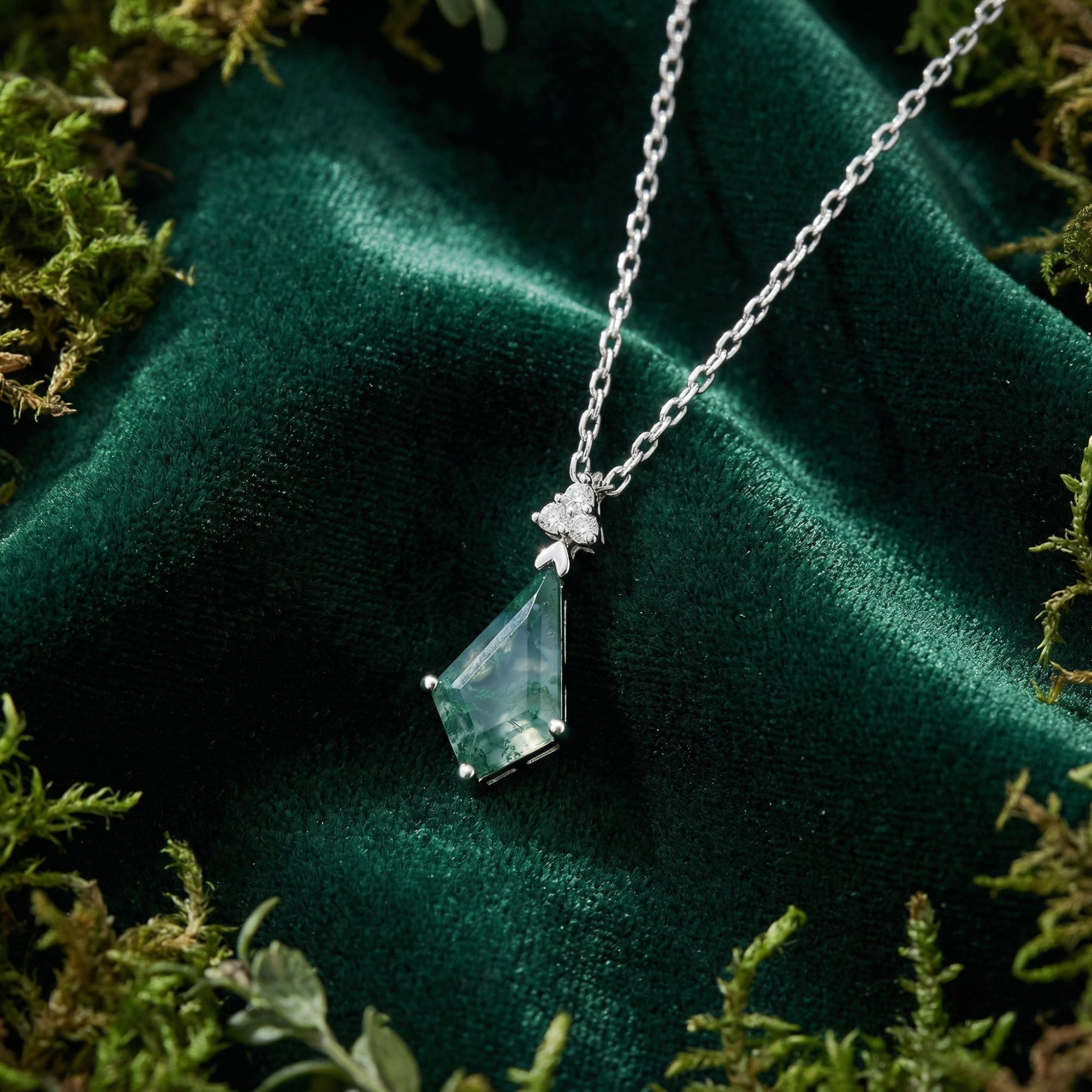 Close-up of a silver necklace with a geometric kite-cut moss agate pendant on a green velvet background.