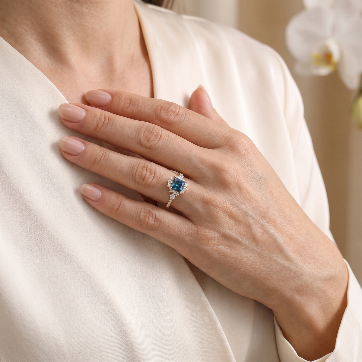 Close-up of a model hand resting on chest wearing a rose gold teal blue moissanite halo ring.