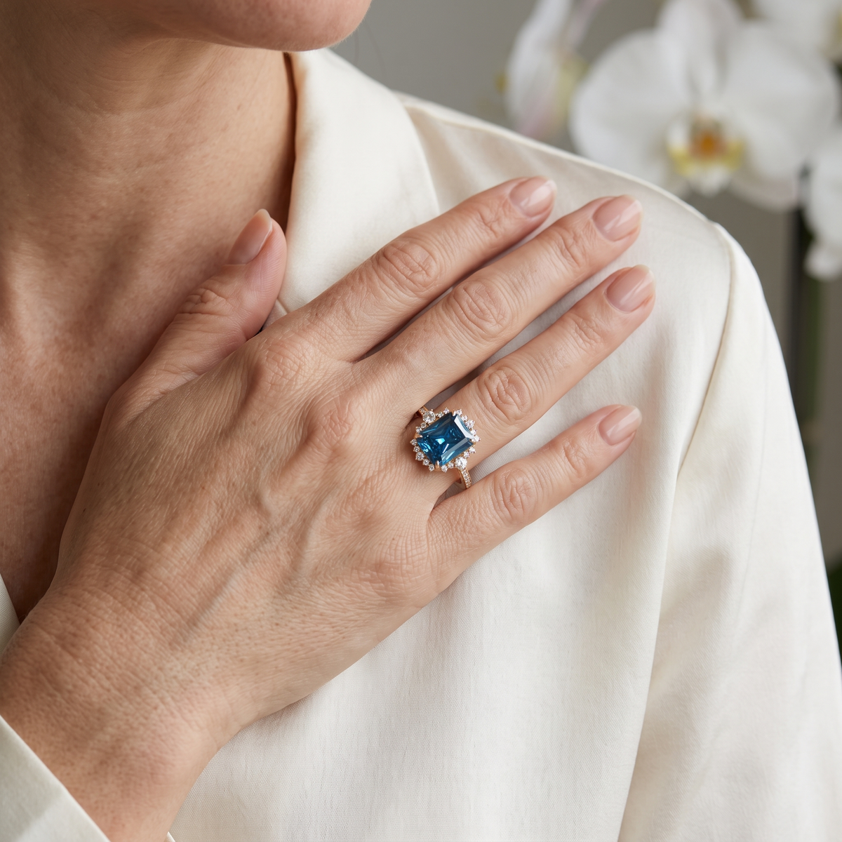 Model wearing an emerald cut blue moissanite halo engagement ring in rose gold resting hand on shoulder.