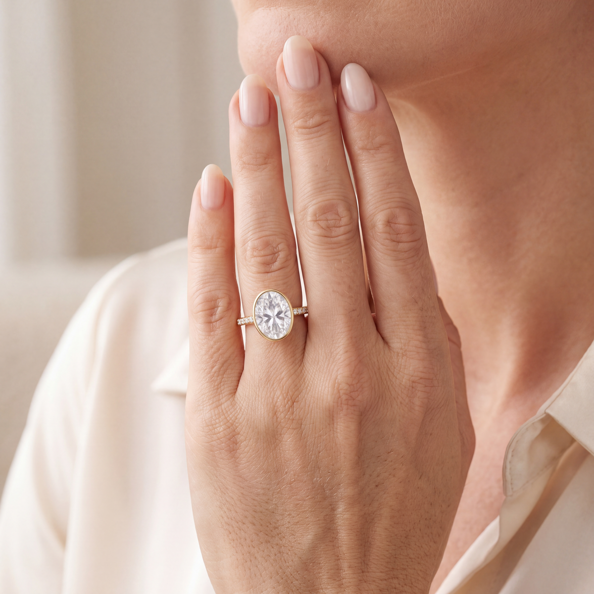 A model holding her hand to her chin showing an oval moissanite bezel engagement ring in yellow gold.