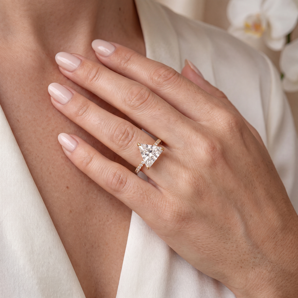 Model wearing a gold trillion-cut moissanite engagement ring with a pavé band against a cream backdrop.