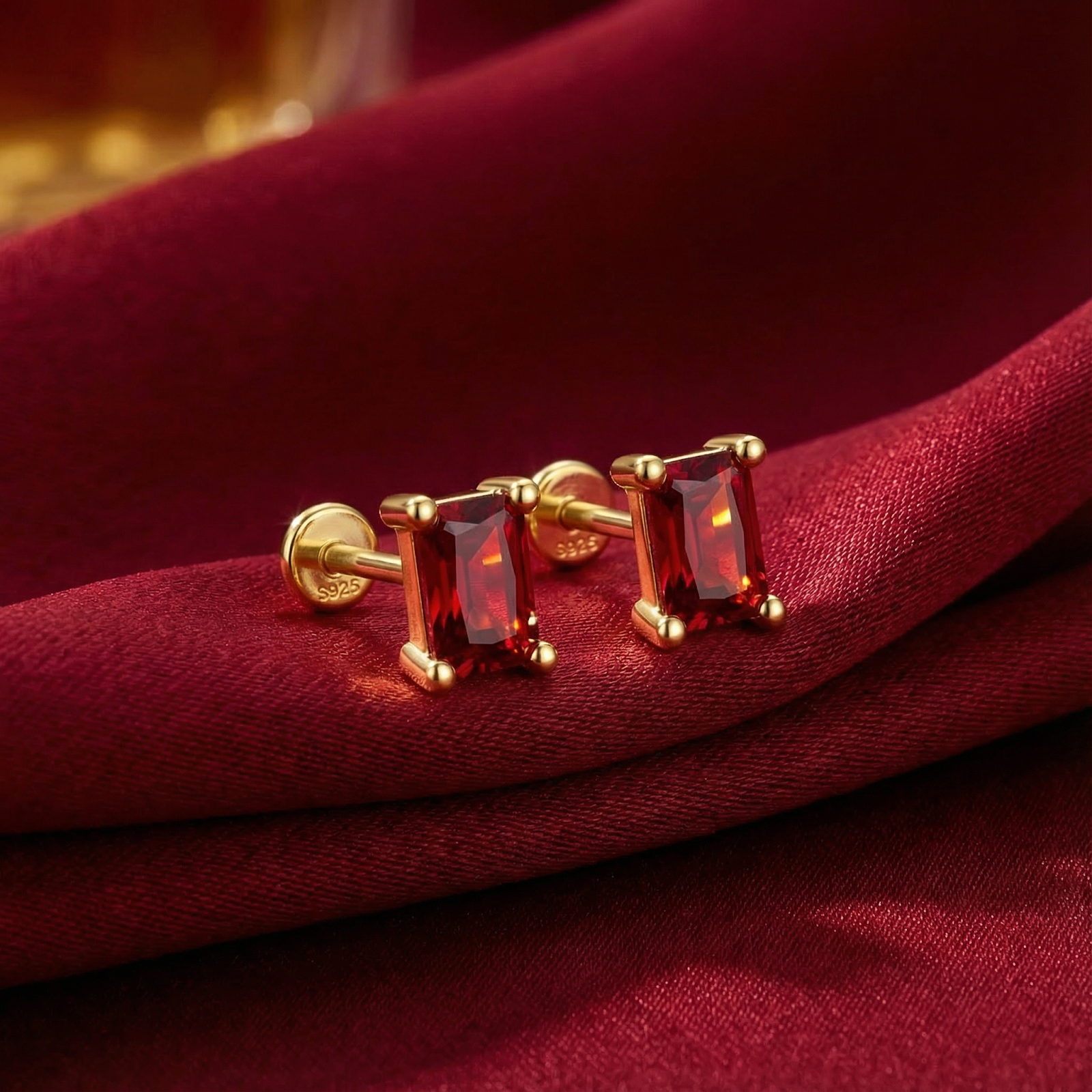 Emerald-cut simulated ruby stud earrings in 18K gold vermeil resting on draped red fabric.