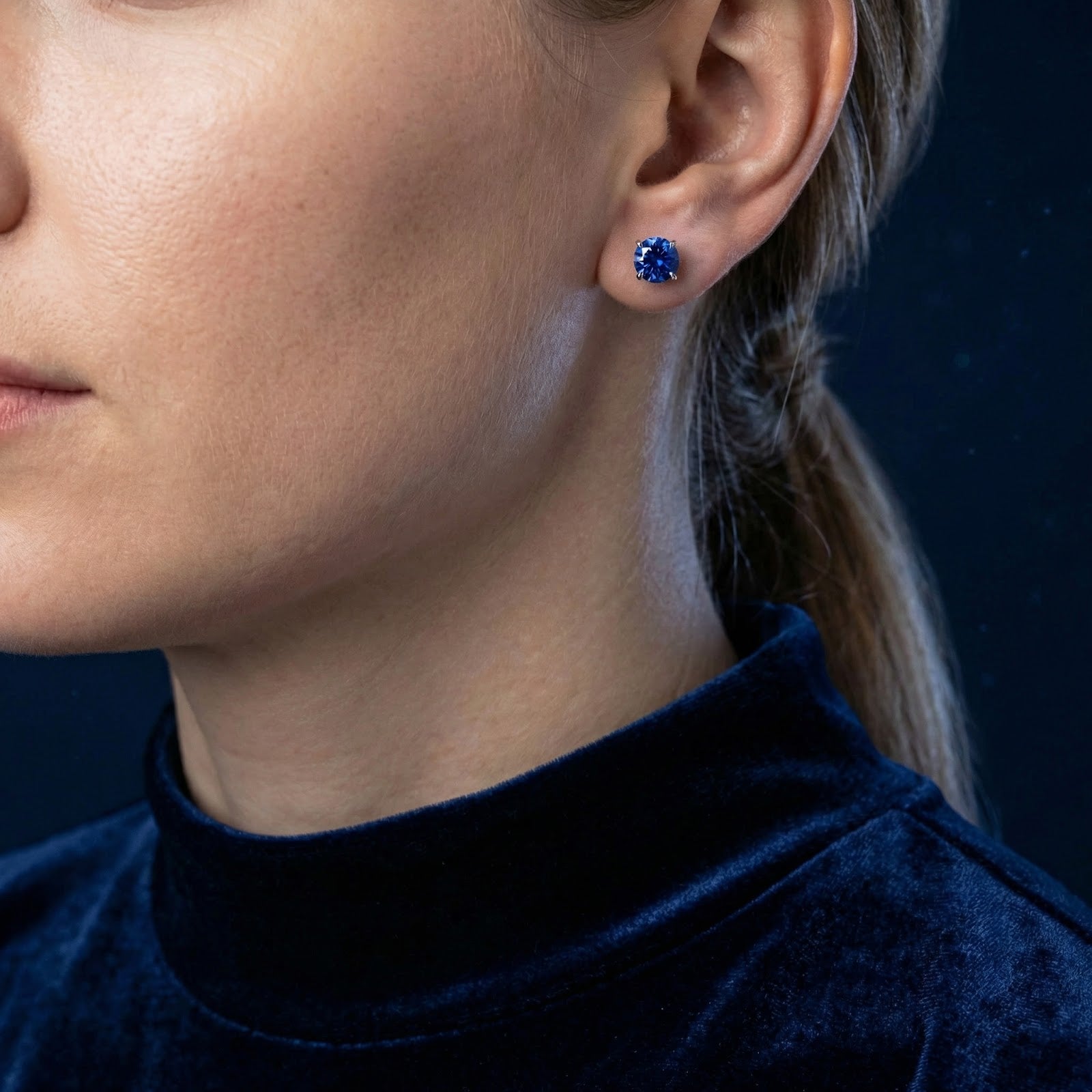 Woman wearing the Esme round blue sapphire stud earrings in sterling silver with a matching blue velvet top.