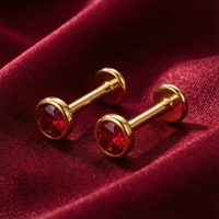 Side profile of gold vermeil labret-style stud earrings with round-cut simulated rubies on red fabric.