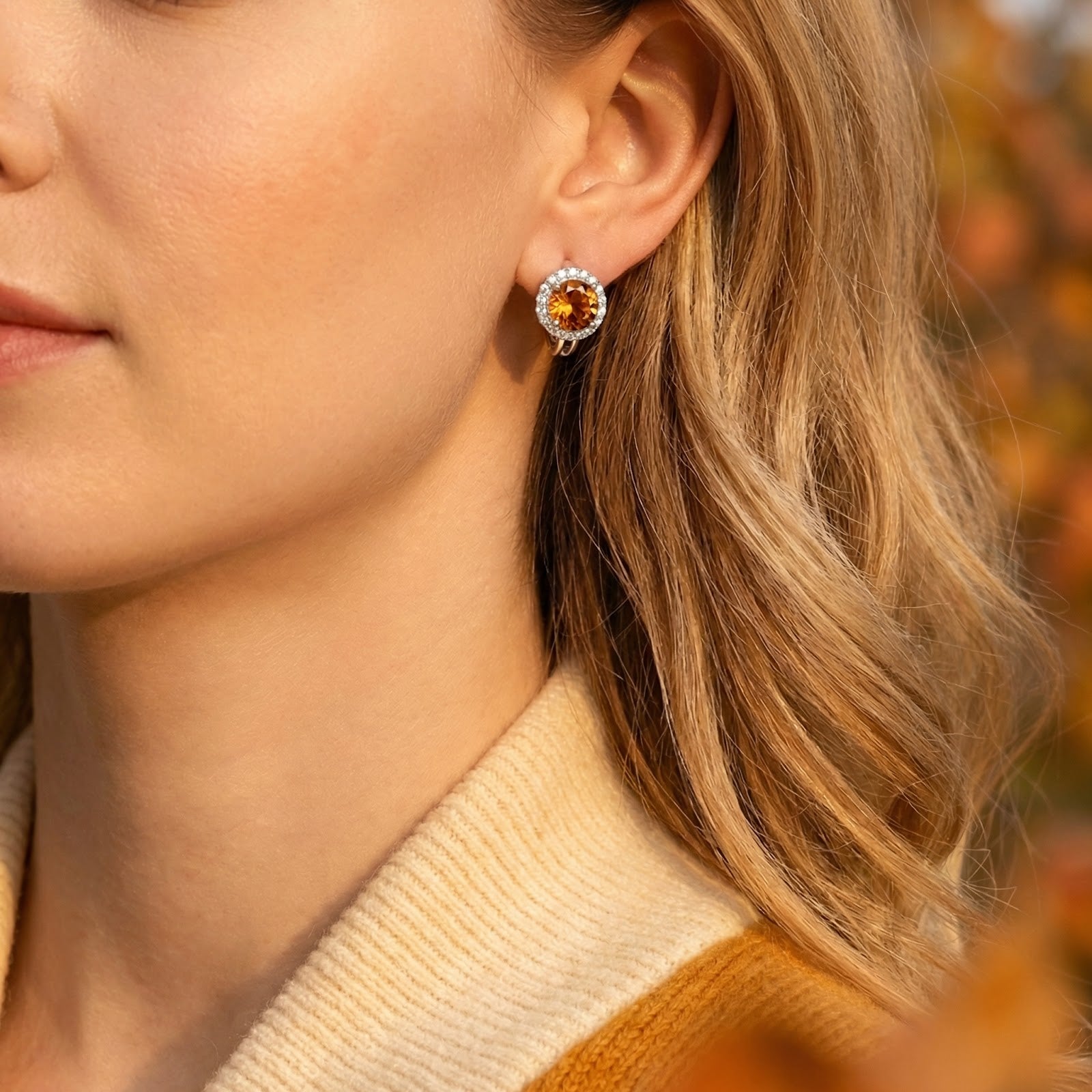 Close-up of a round honey moissanite halo earring worn on a model's ear with a cream knit sweater.