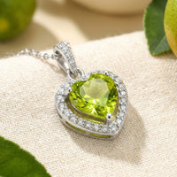 Heart-cut simulated peridot necklace in S925 sterling silver with a halo setting, resting on a beige fabric surface.