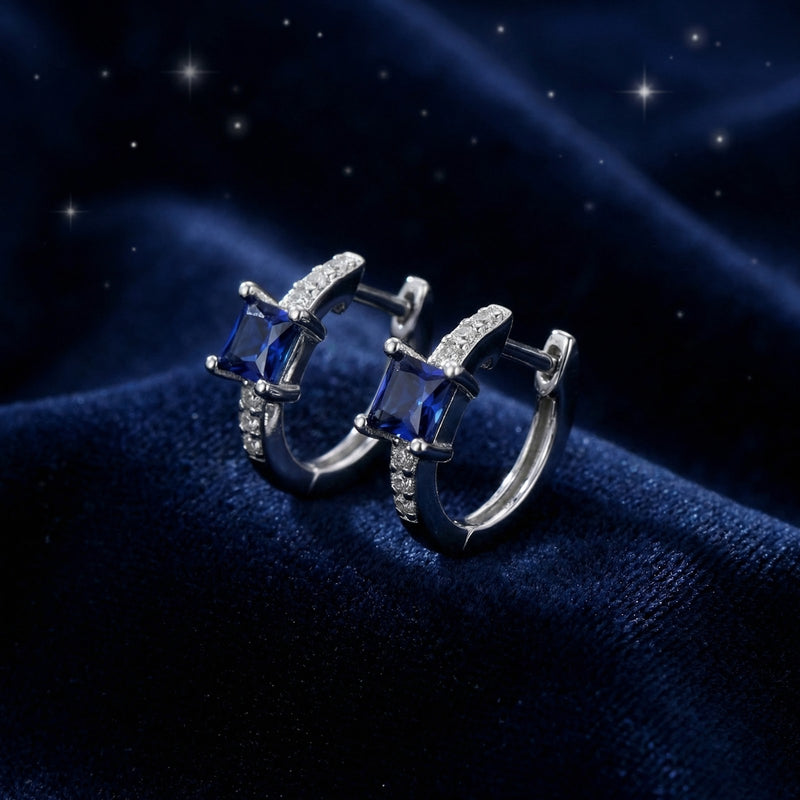 Side view of square-cut blue sapphire huggie earrings in sterling silver on a textured navy background.