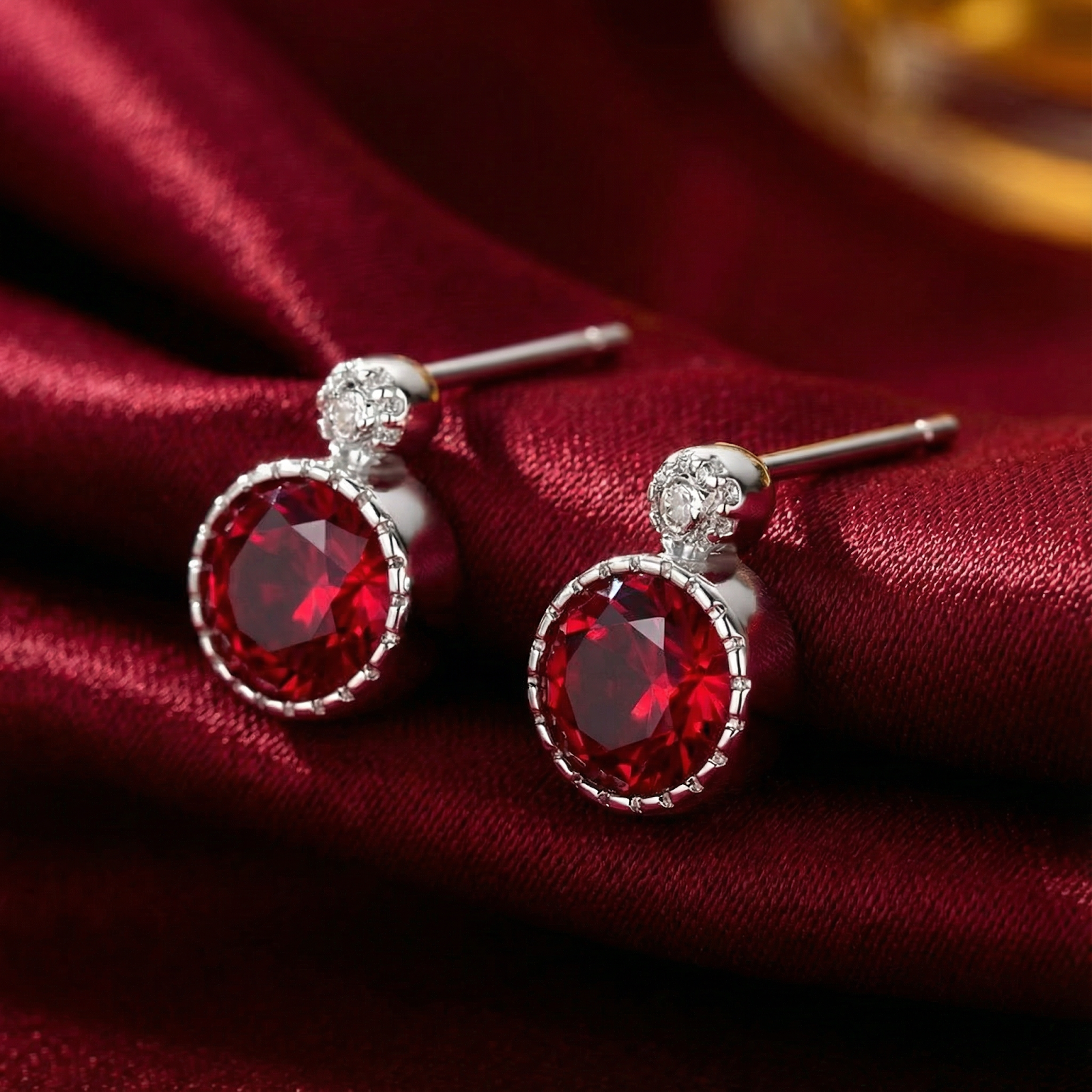 Round cut simulated ruby stud earrings in S925 sterling silver resting on red satin fabric.