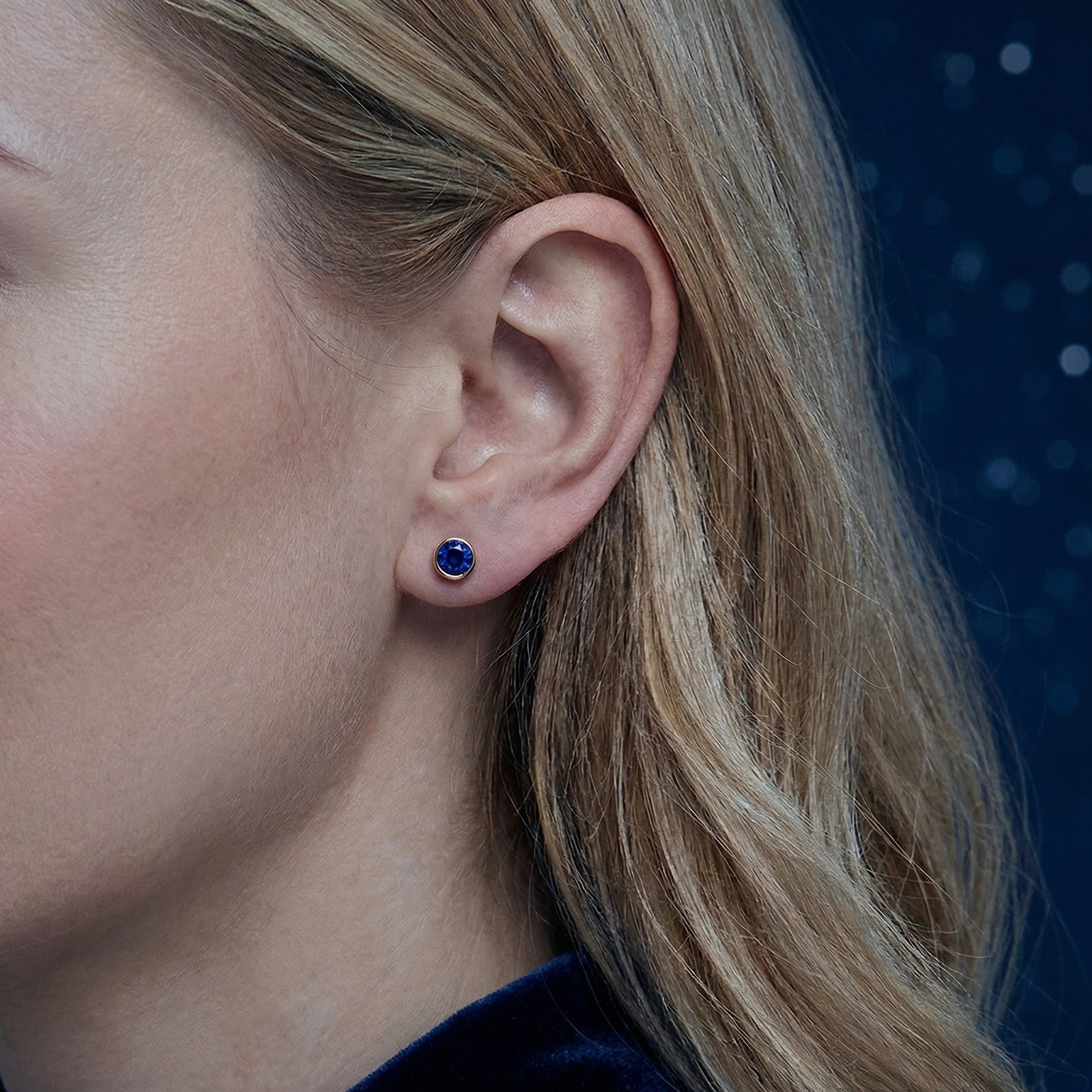Woman wearing a round simulated sapphire bezel stud earring in 18K gold vermeil on her earlobe.