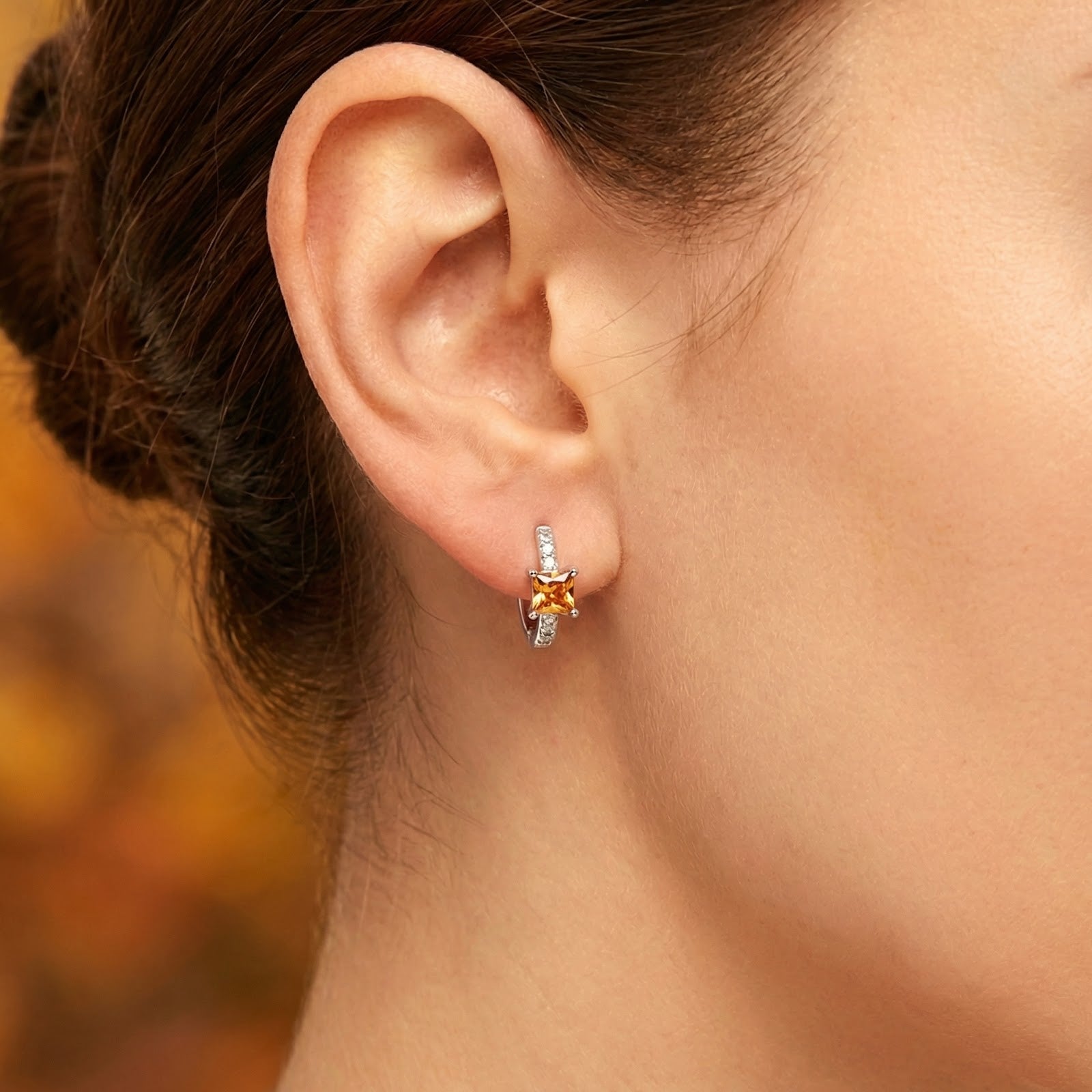 Close-up of a silver huggie earring with a square-cut orange topaz stone worn on a woman's earlobe.