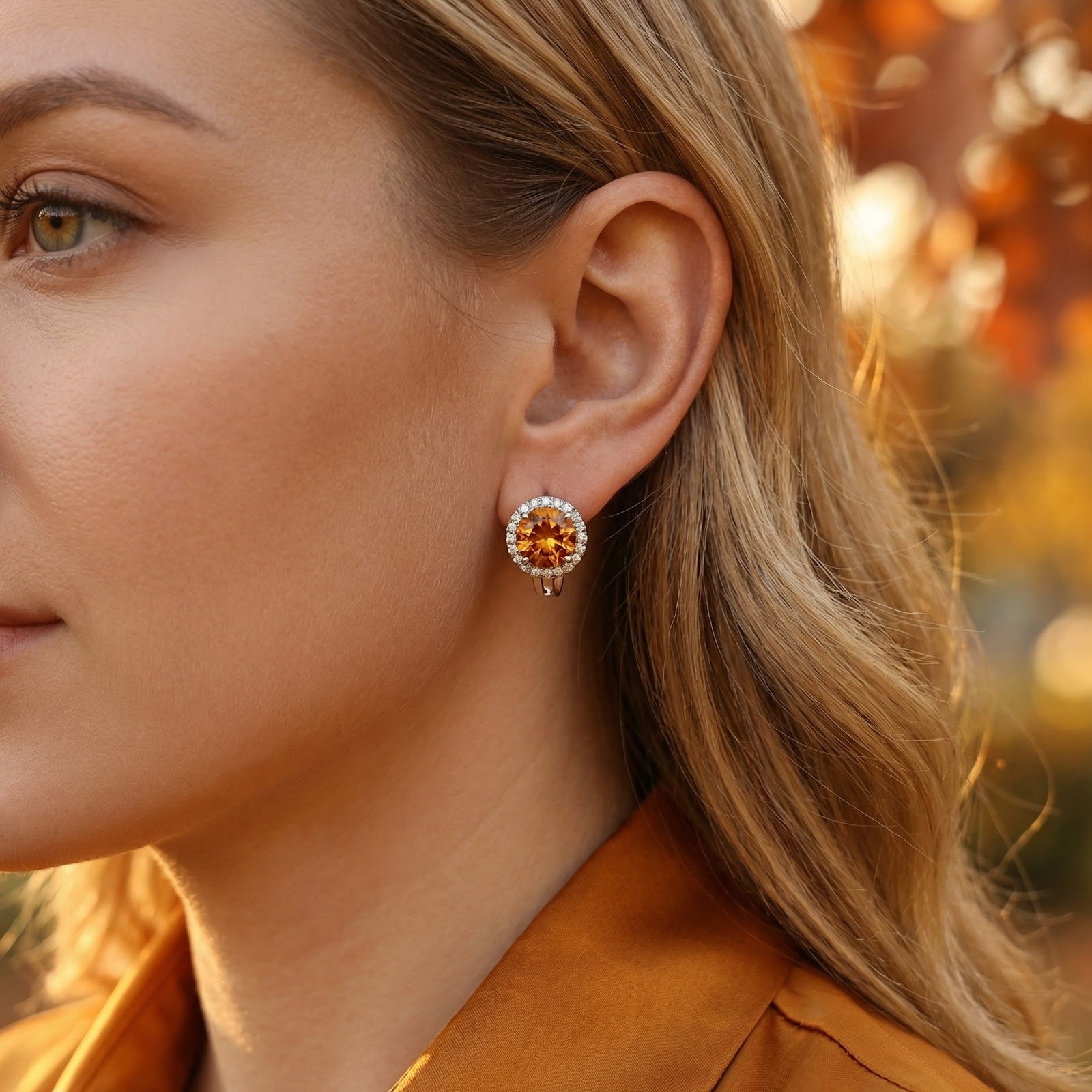 Woman wearing a round honey moissanite halo earring in sterling silver against a blurred autumn backdrop.