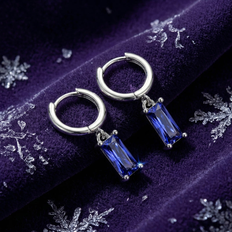 Emerald-cut simulated tanzanite drop earrings in silver-toned metal displayed on purple velvet with snowflakes.