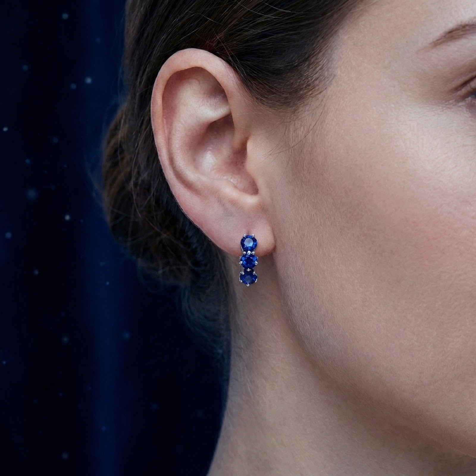 Close-up of a woman wearing a sterling silver star huggie earring with three royal blue simulated sapphires.