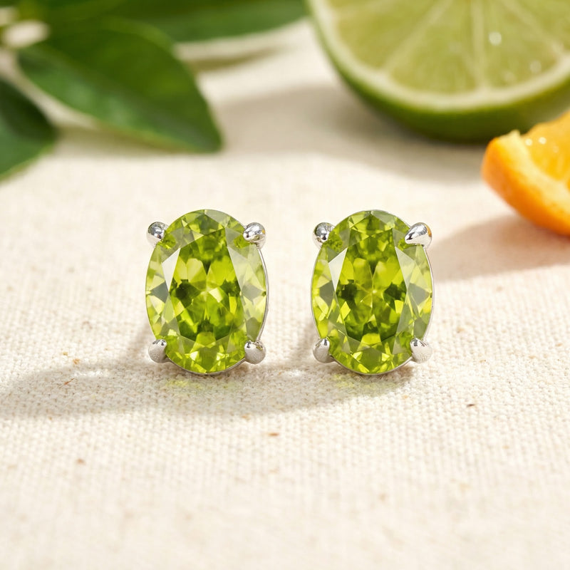 Front view of oval-cut simulated peridot stud earrings in silver 4-prong settings with citrus fruit background.