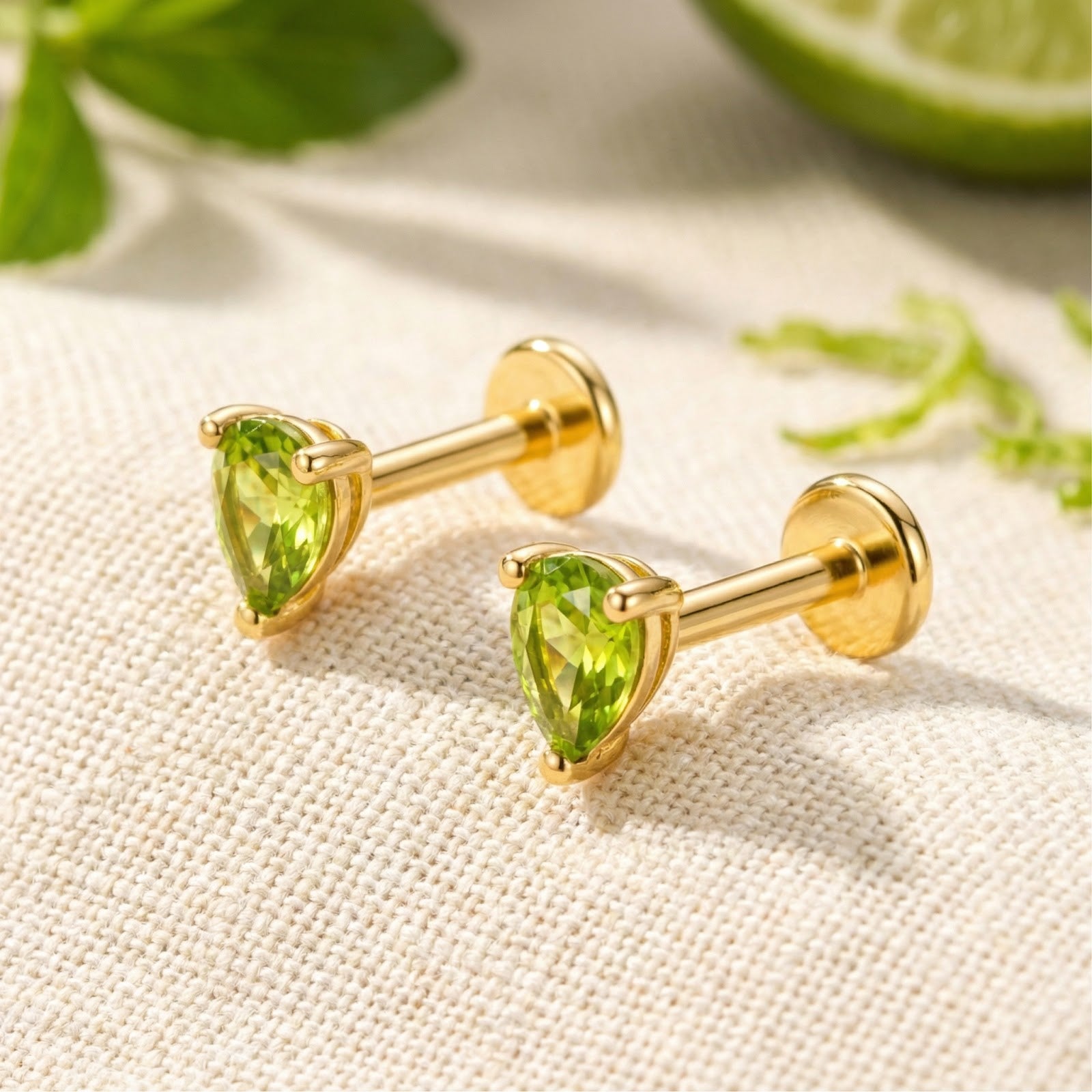 Pear-cut green simulated peridot stud earrings in 18K gold vermeil on a textured fabric lifestyle background.