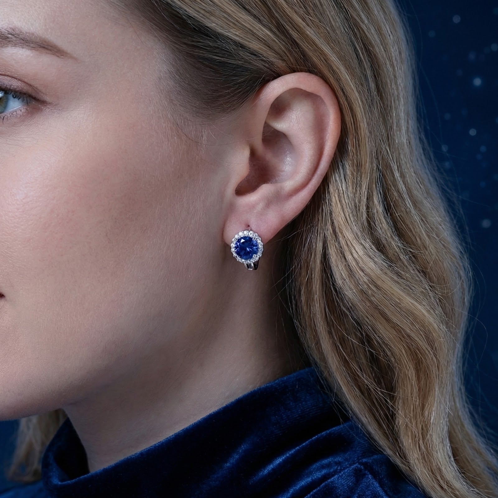 Close-up view of a round blue moissanite halo stud earring in sterling silver worn on a model's ear.