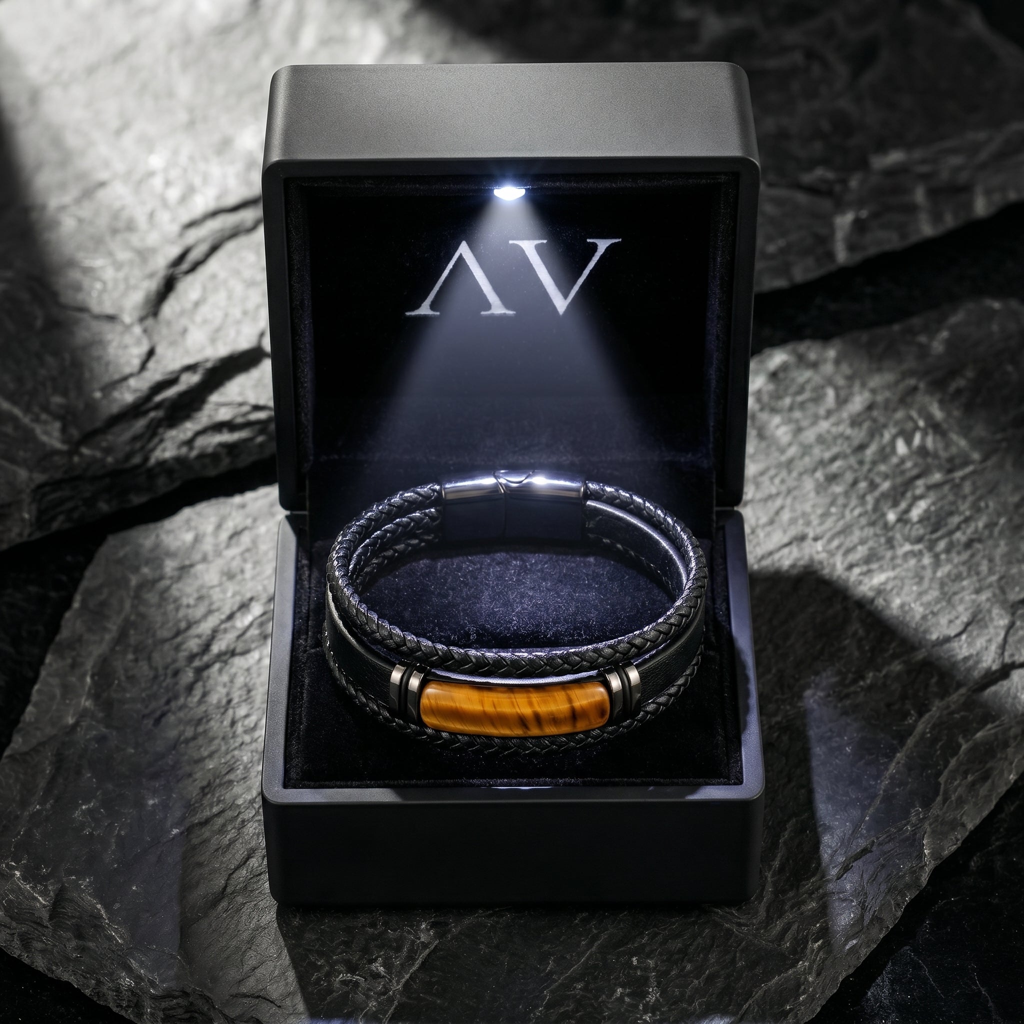 Black braided leather bracelet with tiger's eye stone in a premium black LED-lit gift box on a stone surface.