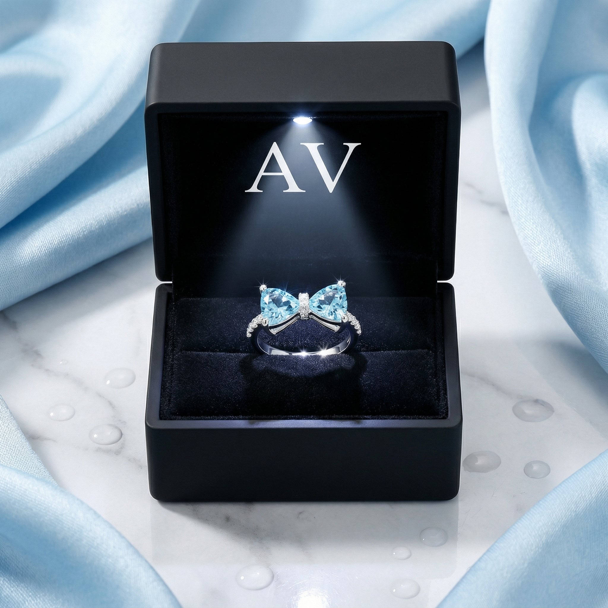 Simulated aquamarine bow ring presented in a black LED-lit jewelry box with AV branding.