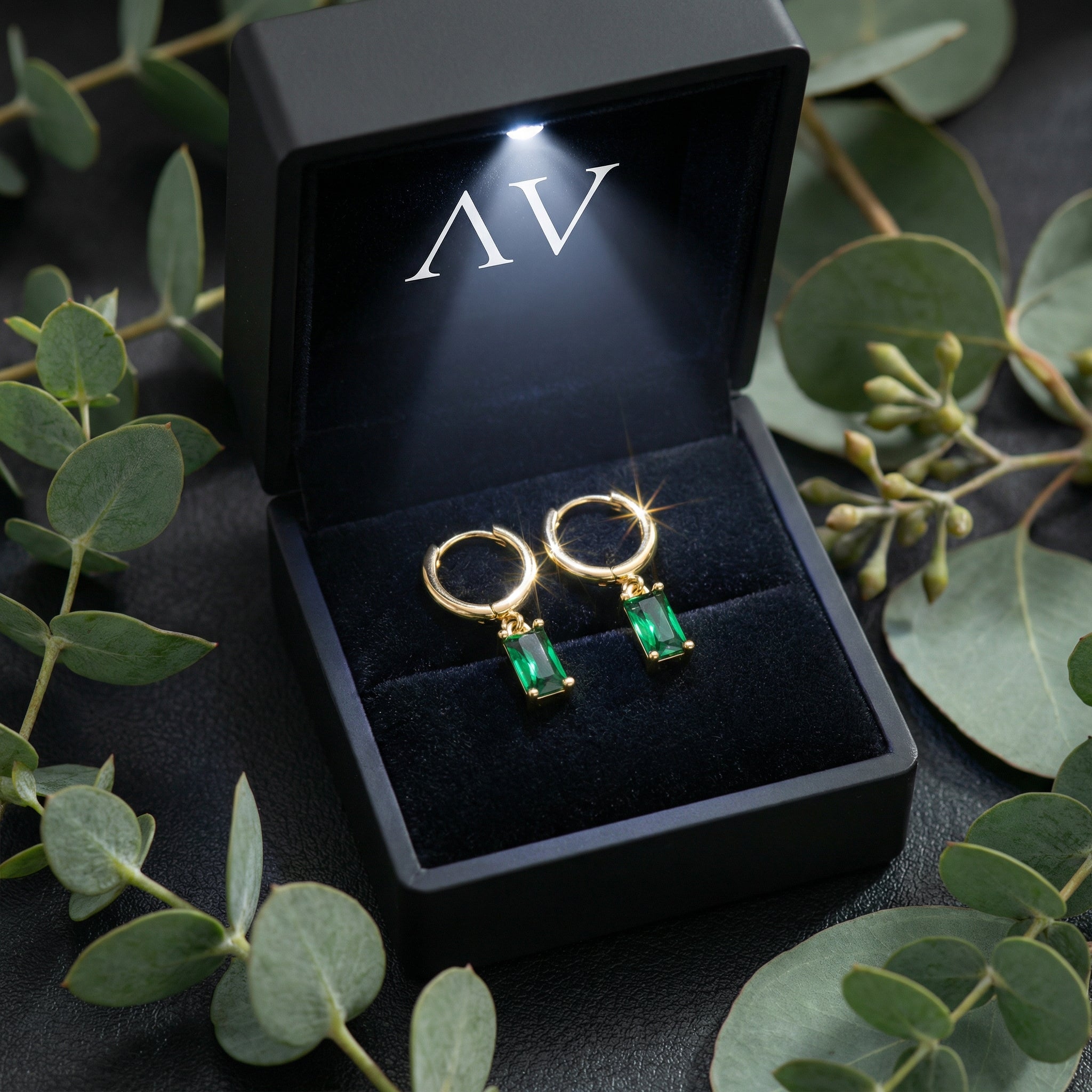 Gold emerald-cut drop earrings in a lighted black gift box styled with eucalyptus leaves on a dark surface.