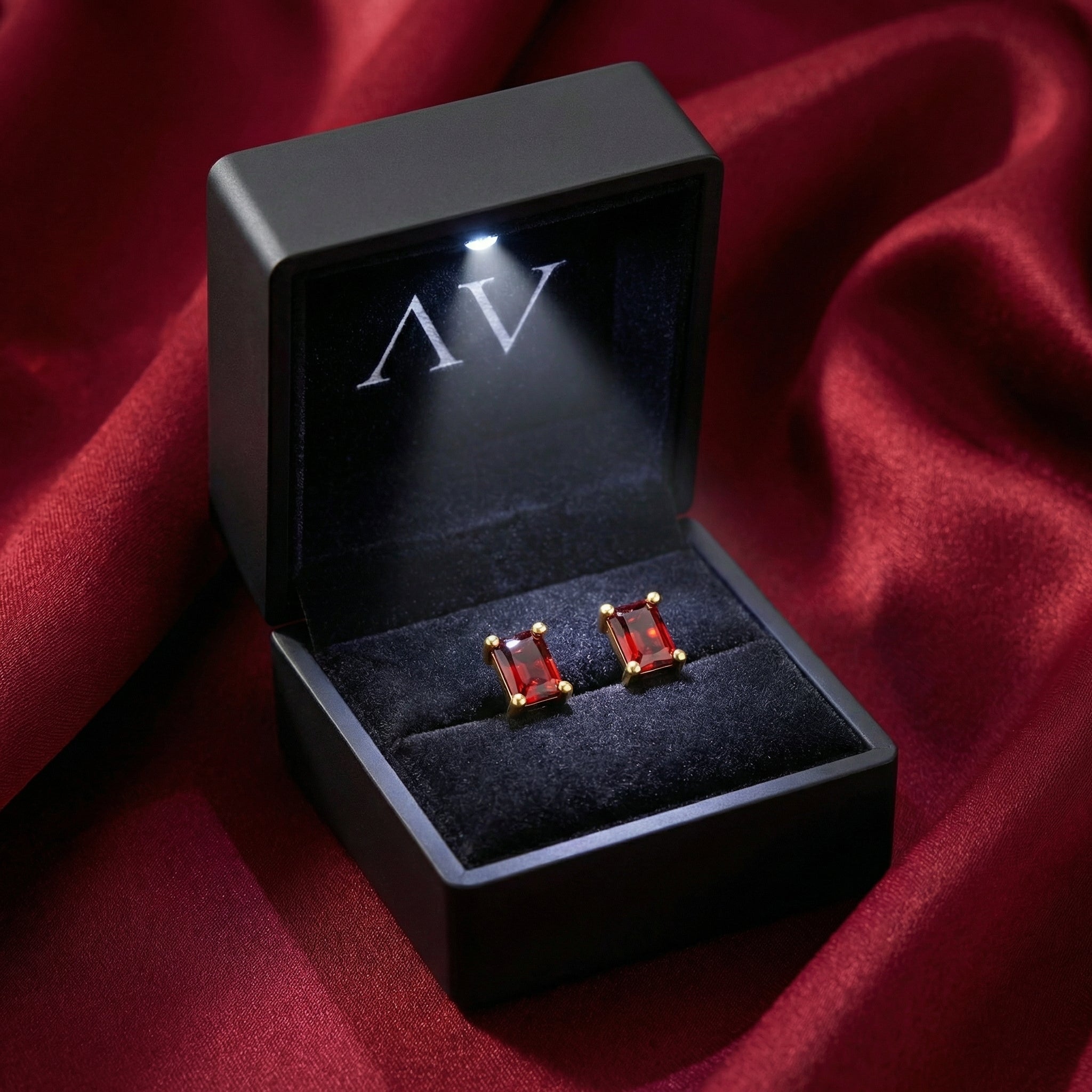 Pair of emerald-cut simulated ruby earrings displayed in a black LED-lit jewelry box on red fabric.
