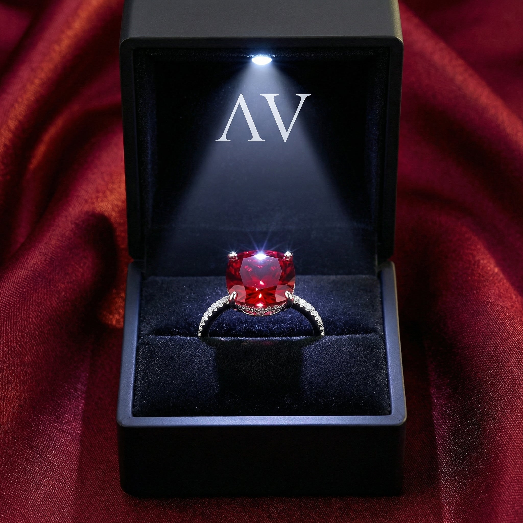 Cushion-cut simulated ruby ring in a black LED-lit presentation box with AV logo on red fabric.