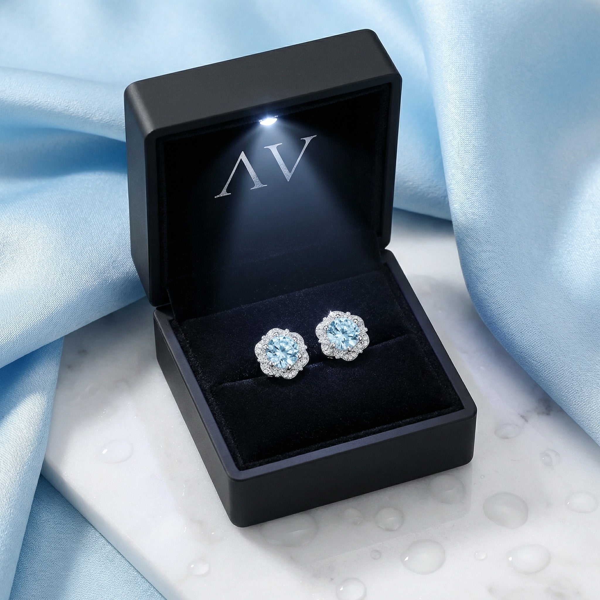 Simulated aquamarine halo stud earrings presented in a black LED-lit jewelry box on blue silk.