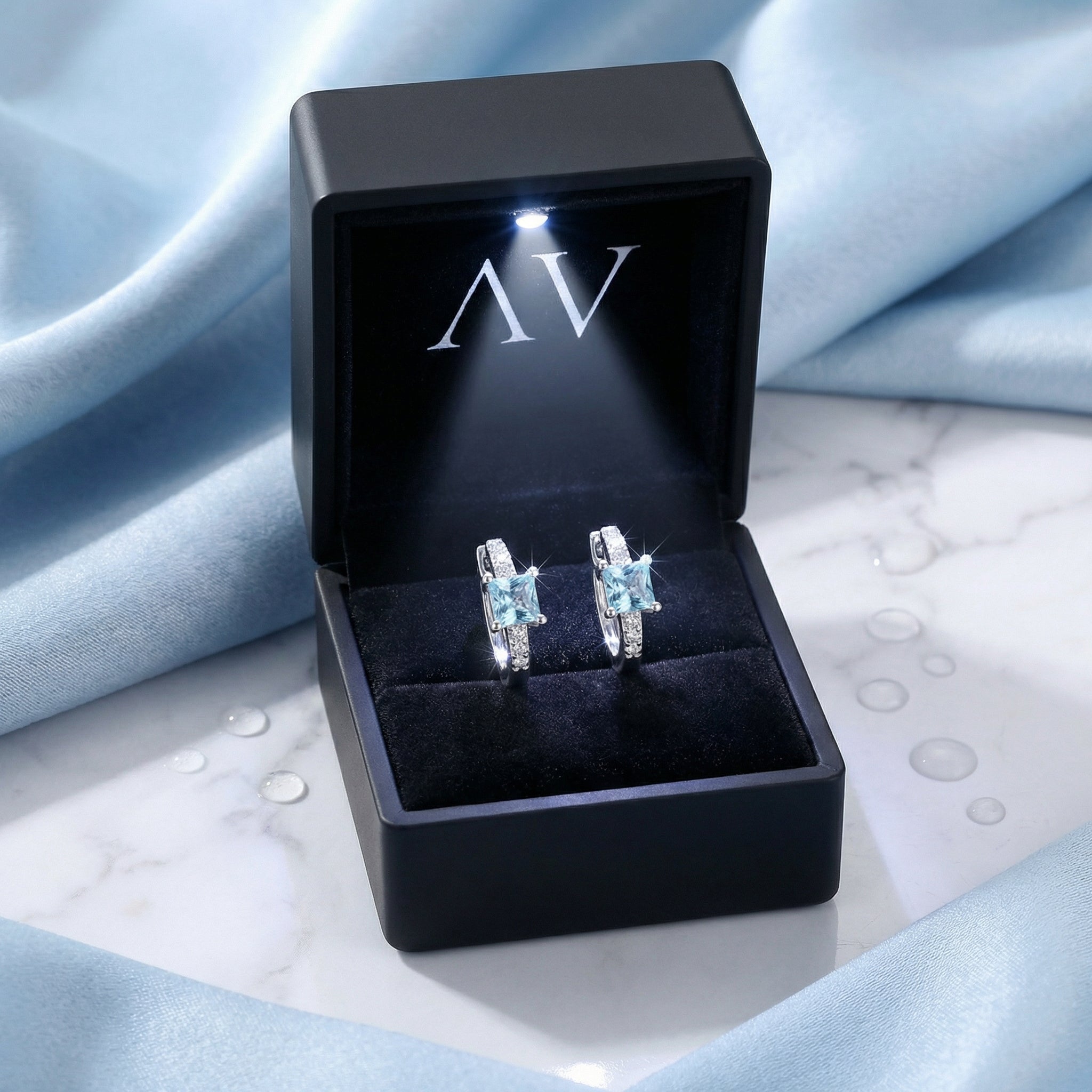 Silver Aira huggie earrings with square simulated aquamarine stones in a black LED-lit jewelry presentation box.