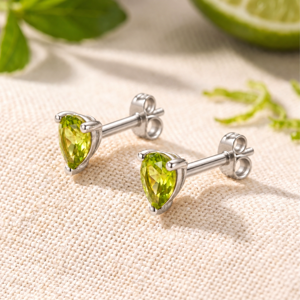 Pair of pear-cut green simulated peridot stud earrings in silver with butterfly backs on textured fabric.