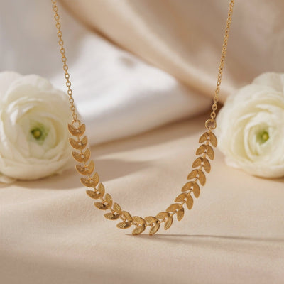Gold-tone leaf necklace arranged on beige fabric with two white flowers.
