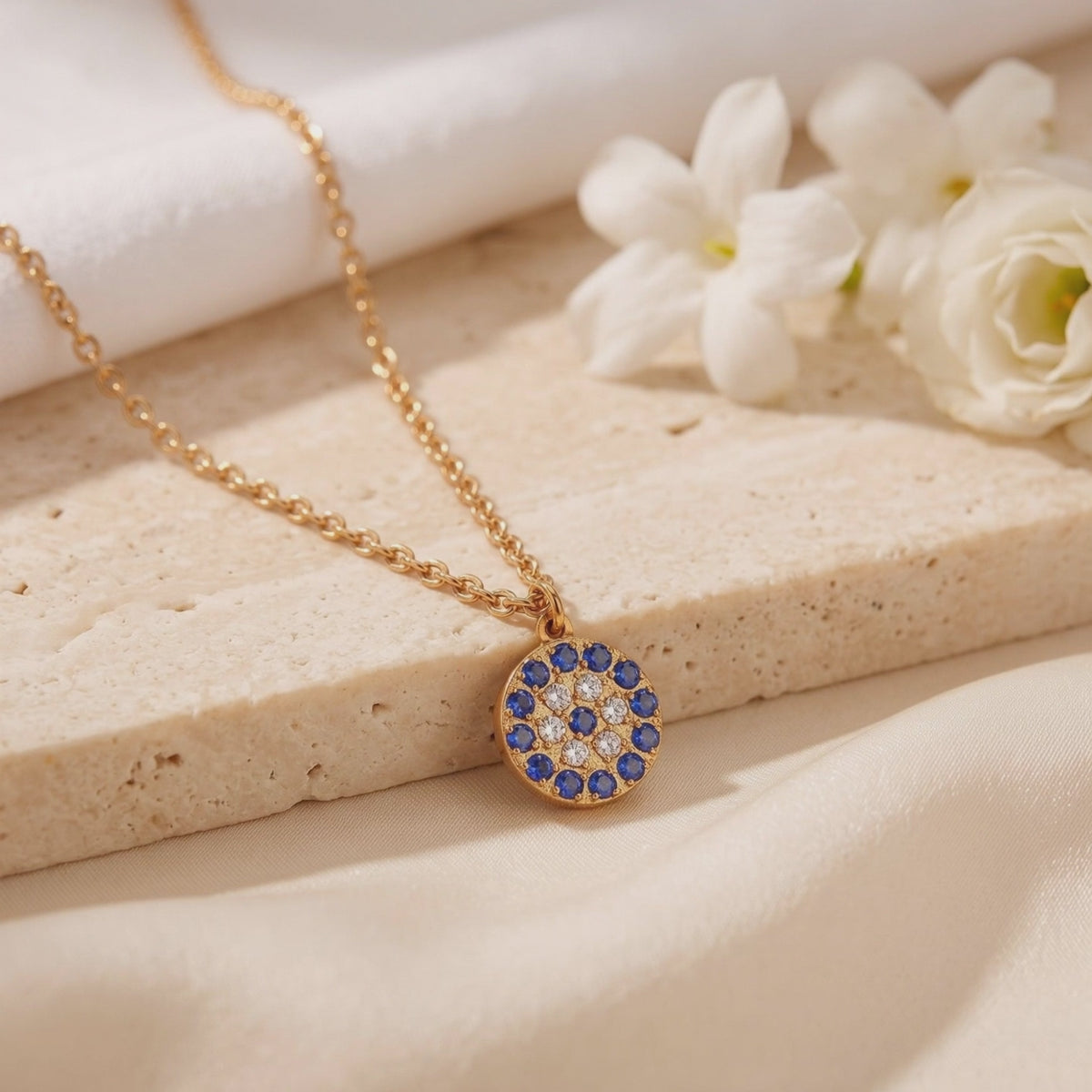 Close-up of a round gold-toned pendant with blue-and-white pavé stones on a pale stone slab beside white flowers.