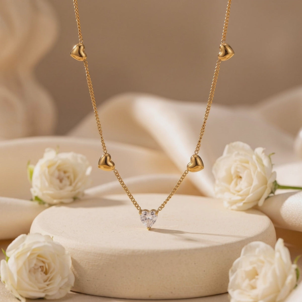 Gold-tone heart station necklace with a clear heart pendant displayed on a beige pedestal among white roses.