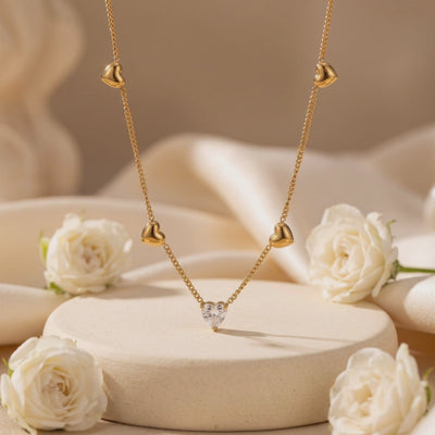 Gold-tone heart station necklace with a clear heart pendant displayed on a beige pedestal among white roses.
