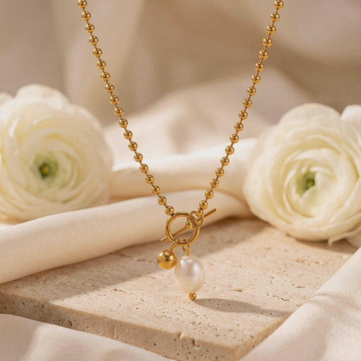 Gold-toned bead chain necklace displayed on a stone block with a toggle clasp, pearl, and sphere charm.