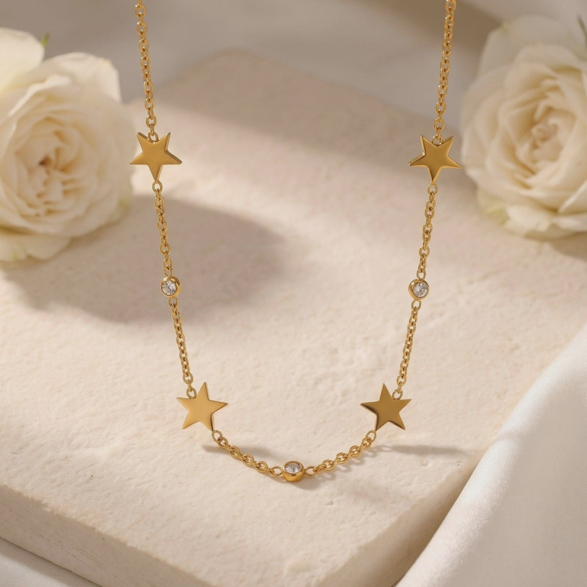 Close-up of a gold-tone star-charm necklace with round simulated diamond accents on a textured block among white roses.