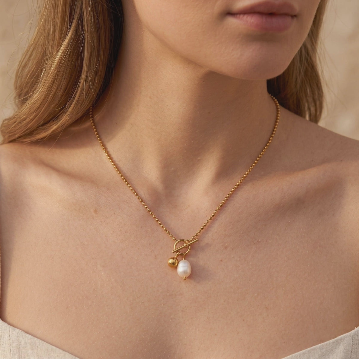 Gold-toned bead chain necklace worn on a model, with a front toggle clasp, pearl, and sphere charm.