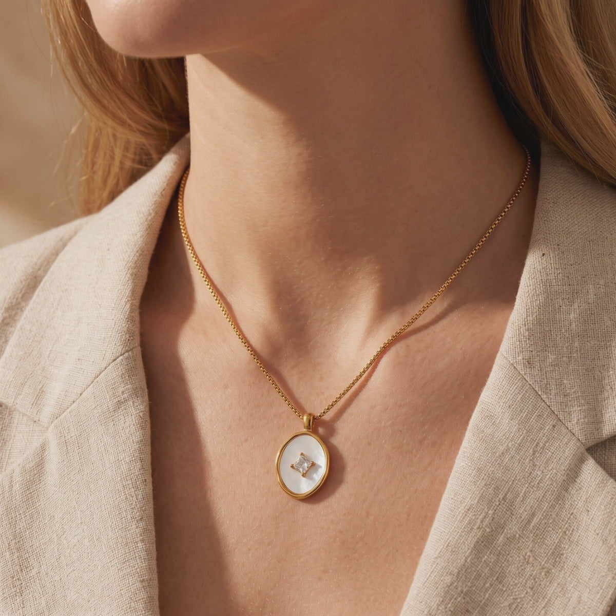 Gold-tone necklace with an oval mother-of-pearl pendant and square center stone, worn over a beige blazer.